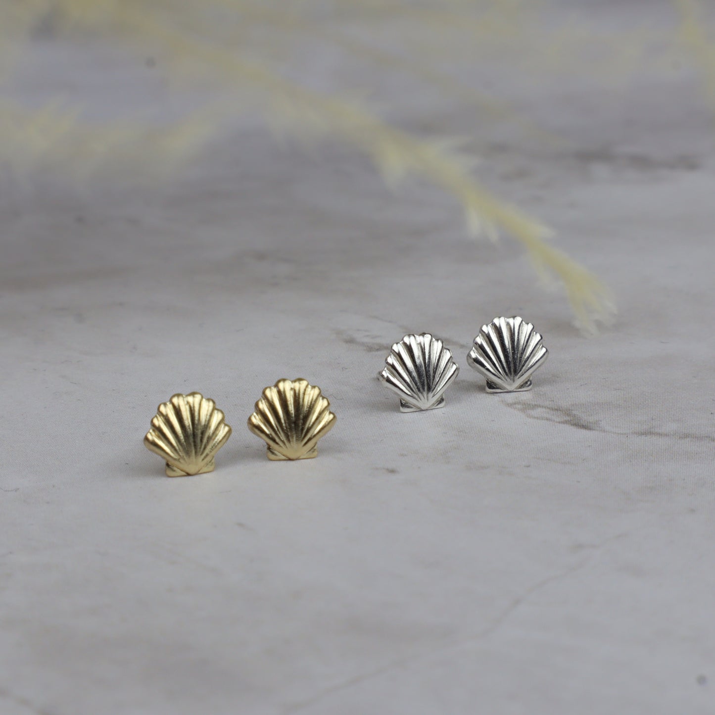 Four scallop-shaped stud earrings, two in yellow gold and two in silver, arranged on a light gray stone surface with soft blurred dried grass in the background.