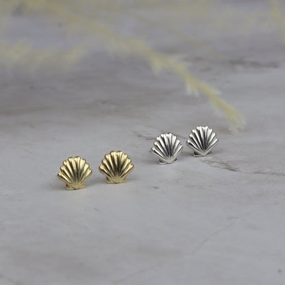 Four scallop-shaped stud earrings, two in yellow gold and two in silver, arranged on a light gray stone surface with soft blurred dried grass in the background.