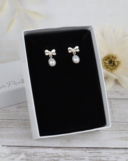 A pair of silver bow stud earrings with white pearl drops, presented in an open white gift box with a black velvet insert on a light wood surface.