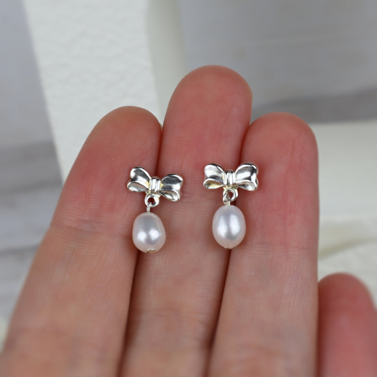 A pair of silver bow stud earrings with dangling baroque pearls, shown resting on a person's fingers.