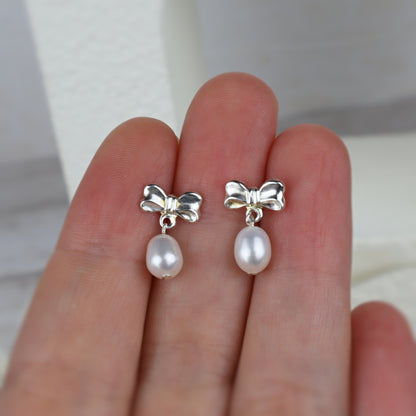 A pair of silver bow stud earrings with dangling baroque pearls, shown resting on a person's fingers.