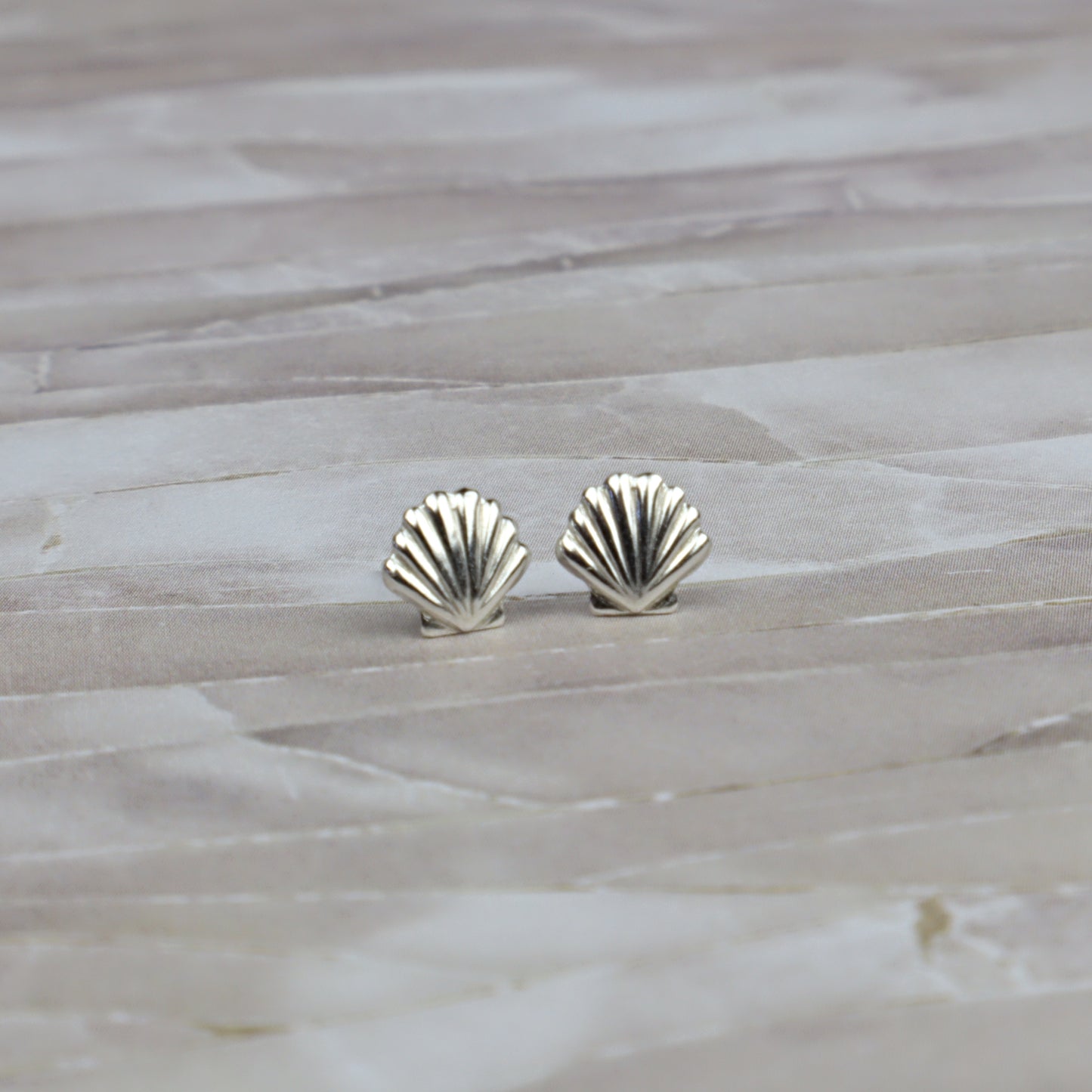 A pair of small silver stud earrings shaped like scallop shells, resting on a light beige stone surface.