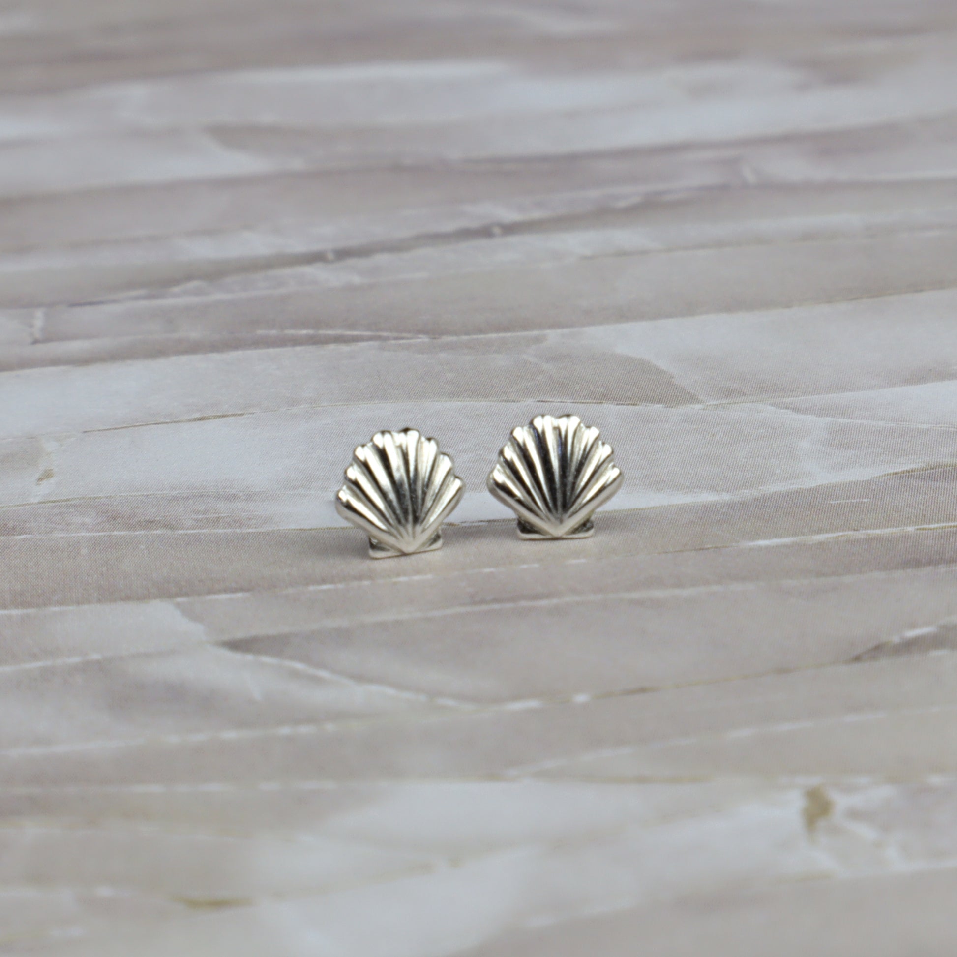 A pair of small silver stud earrings shaped like scallop shells, resting on a light beige stone surface.