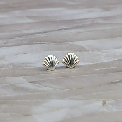 A pair of small silver stud earrings shaped like scallop shells, resting on a light beige stone surface.