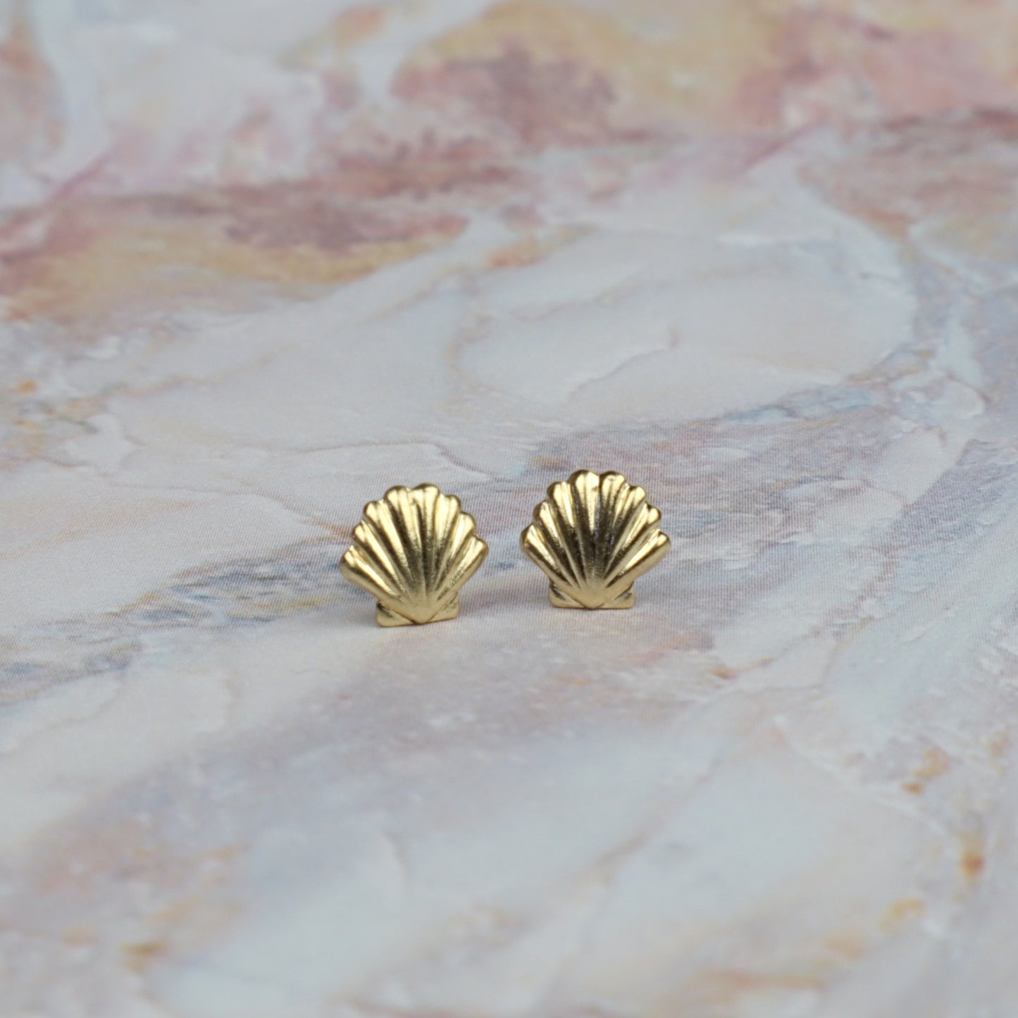 A pair of small gold shell-shaped stud earrings resting on a light pink and white marbled surface.
