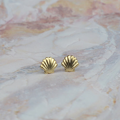 A pair of small gold shell-shaped stud earrings resting on a light pink and white marbled surface.
