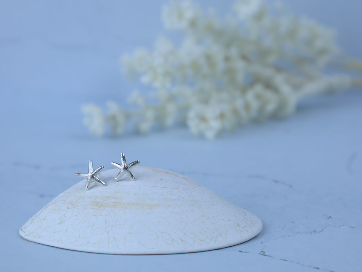 A pair of silver starfish stud earrings displayed on a white ceramic dish against a light blue background with blurred white flowers.