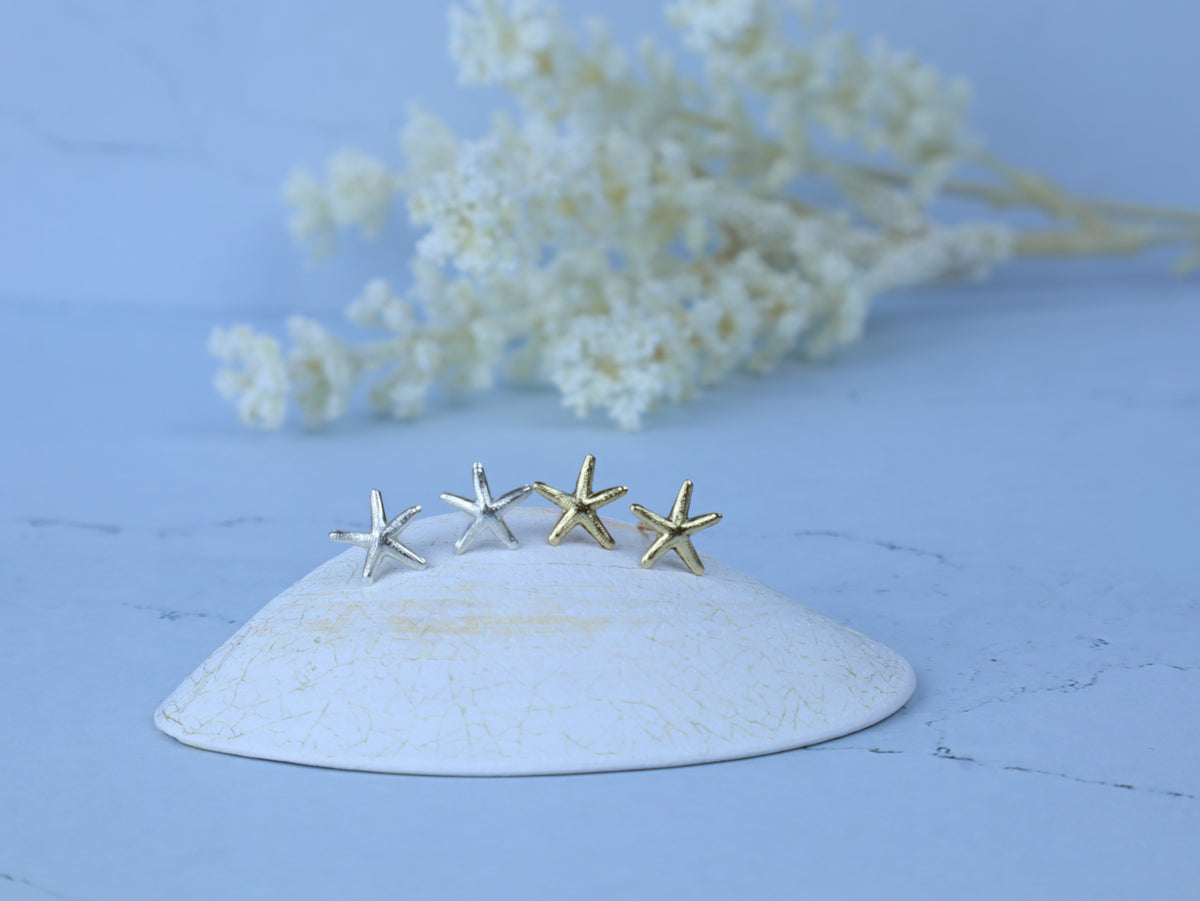 Four small starfish-shaped earrings in silver and gold finishes displayed on a white ceramic dish against a light blue background with blurred white flowers.