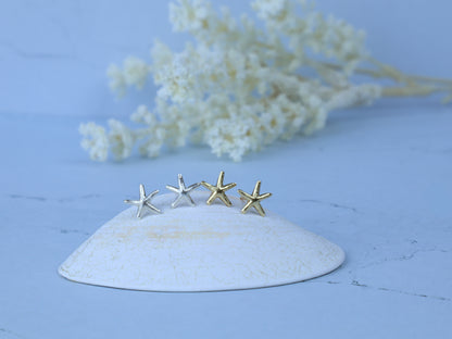 Four small starfish-shaped earrings in silver and gold finishes displayed on a white ceramic dish against a light blue background with blurred white flowers.