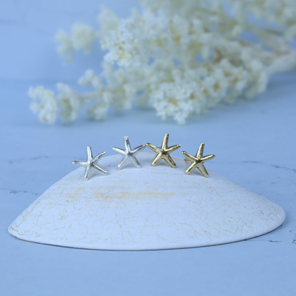 Four small starfish-shaped stud earrings in silver and gold tones displayed on a white ceramic dish against a soft blue background with blurred white flowers.