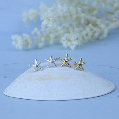 Four small starfish-shaped stud earrings in silver and gold tones displayed on a white ceramic dish against a soft blue background with blurred white flowers.