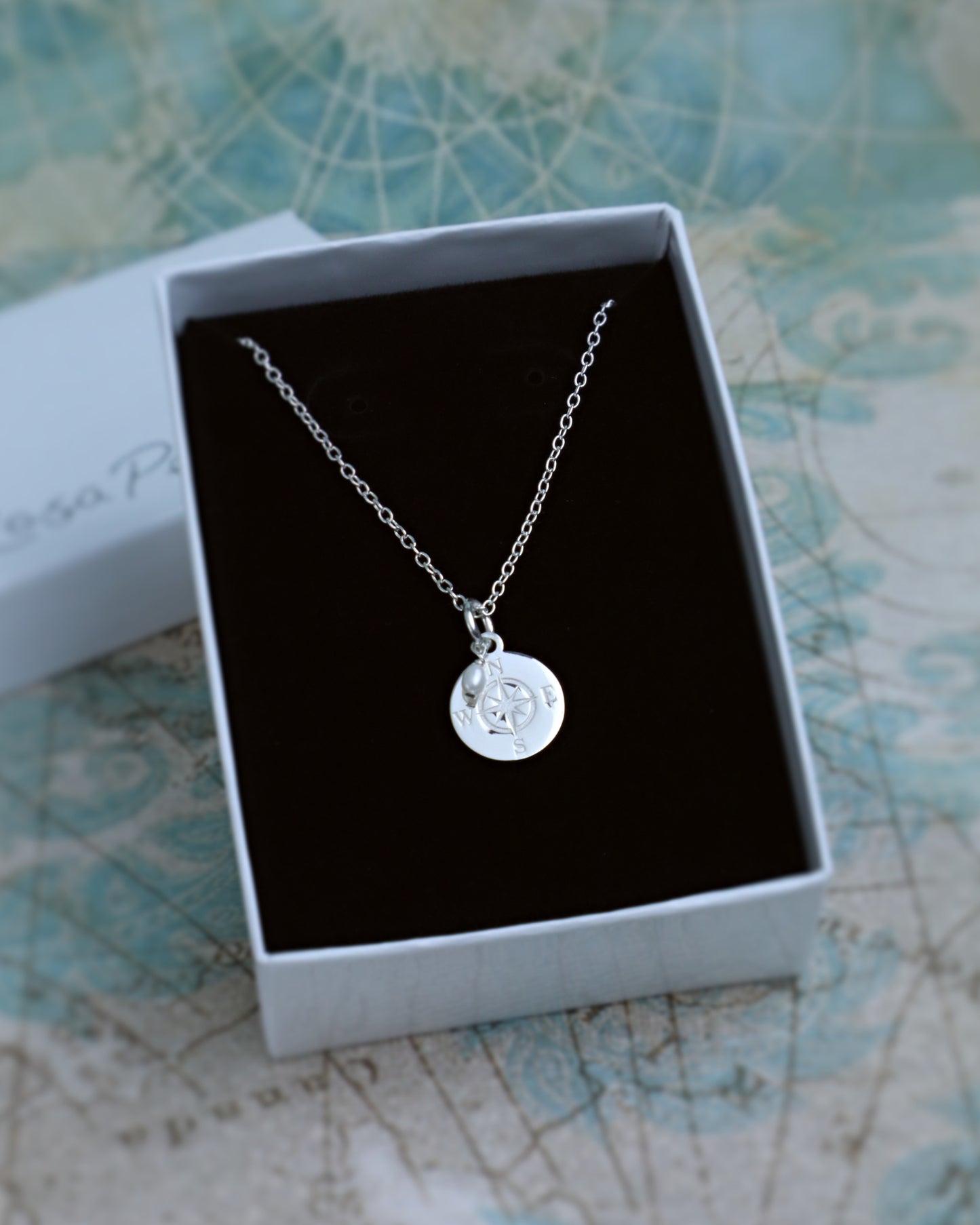 A silver necklace with a circular engraved pendant displayed inside an open white gift box with a black insert, set against a light blue patterned background.
