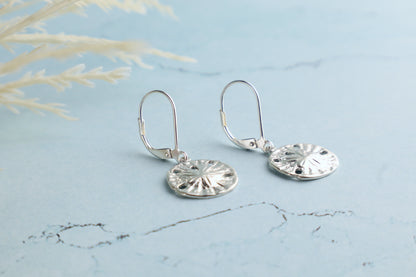 Silver sand dollar earrings on a light blue marble surface with grass in the background