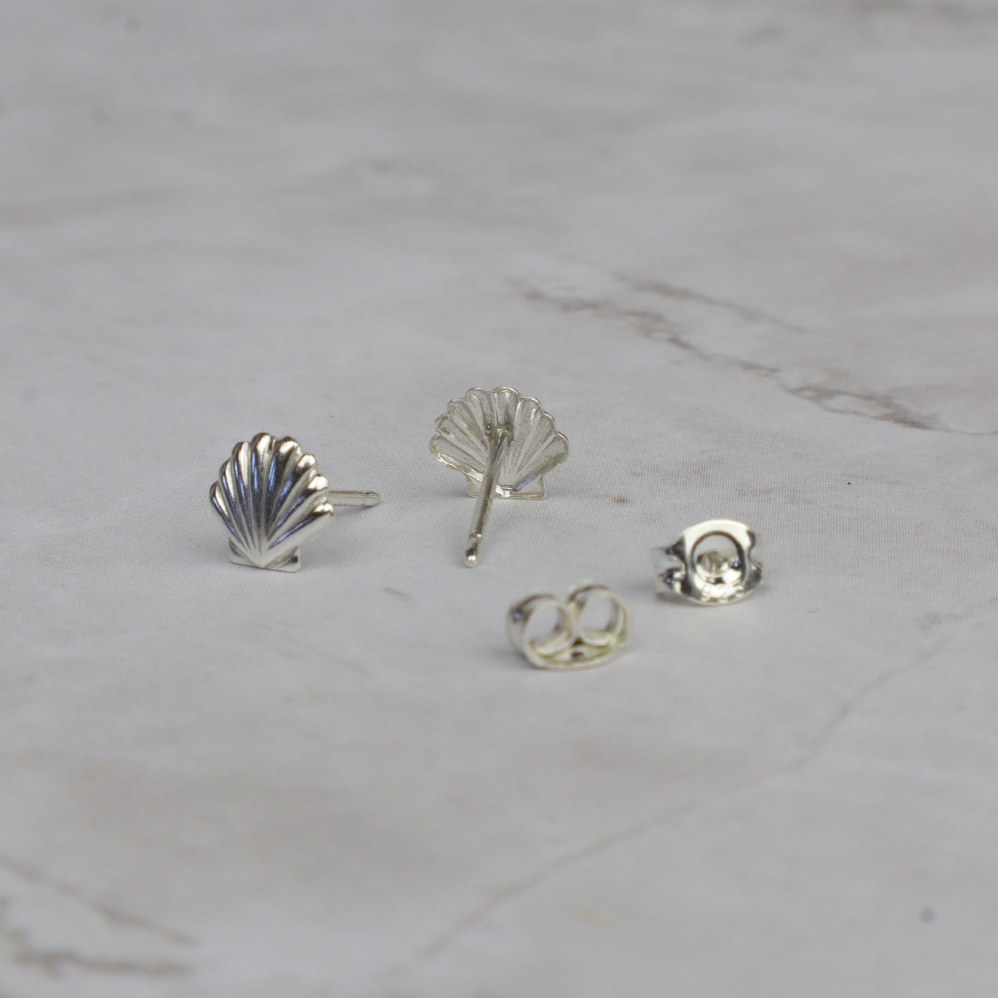 Four silver-colored jewelry components, including two shell-shaped stud earrings, one leaf-shaped stud, and two butterfly backings, arranged on a light gray marble surface.