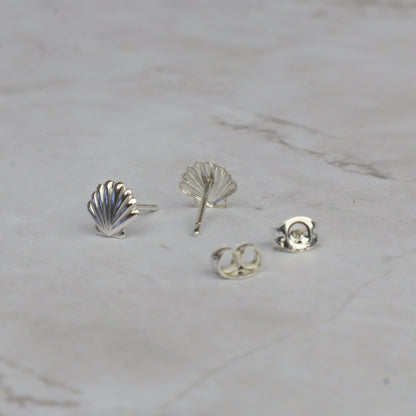 Four silver-colored jewelry components, including two shell-shaped stud earrings, one leaf-shaped stud, and two butterfly backings, arranged on a light gray marble surface.