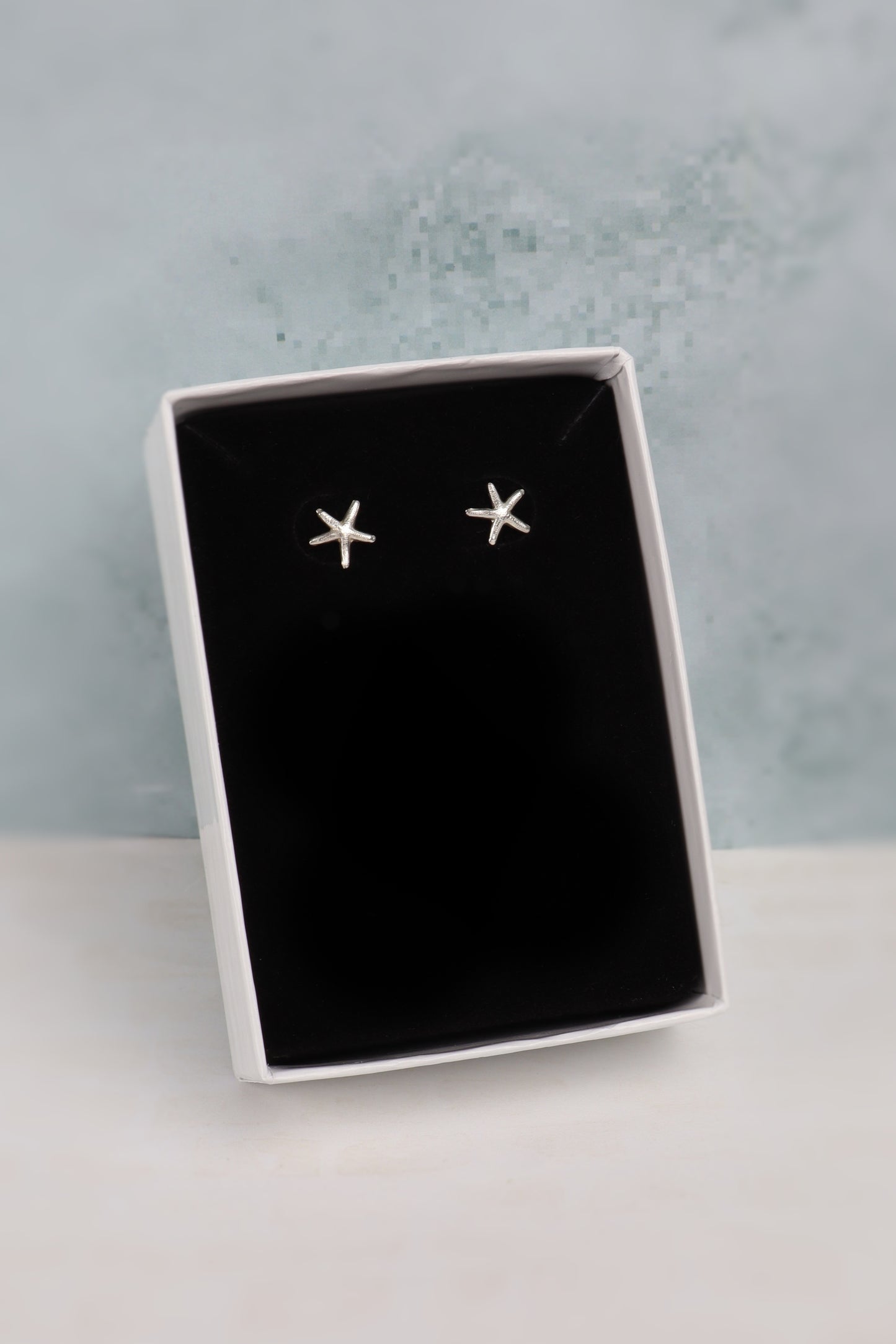 A pair of small silver star-shaped stud earrings displayed on a black insert inside an open white gift box, set against a pale blue textured background.