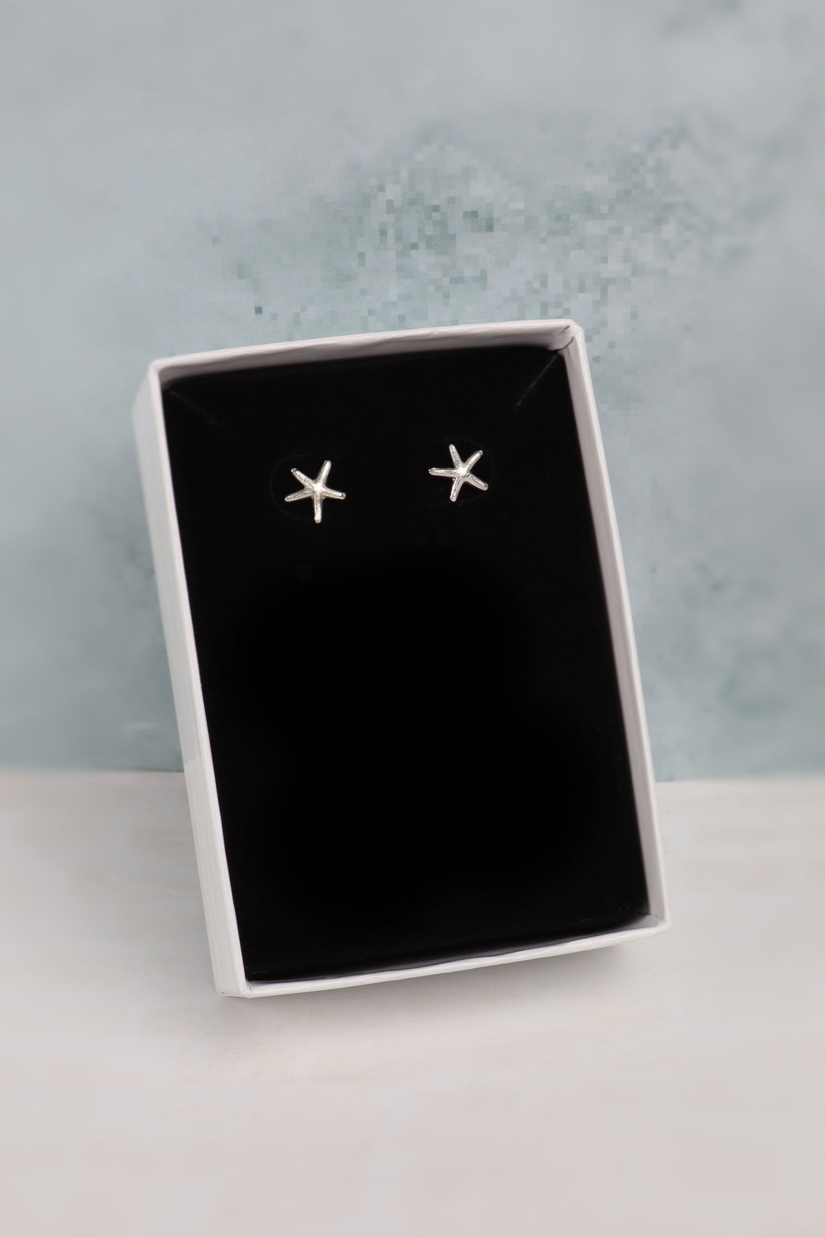 A pair of small silver star-shaped stud earrings displayed on a black insert inside an open white gift box, set against a pale blue textured background.