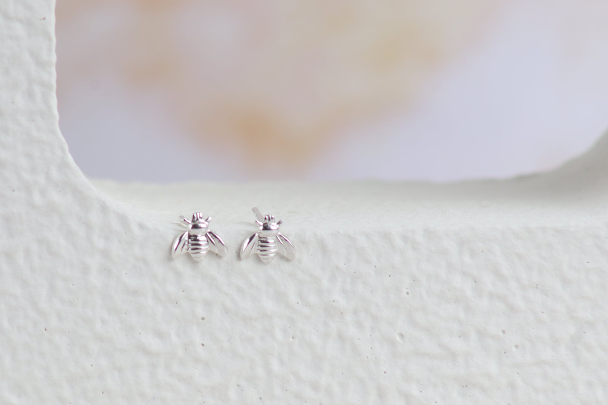 A pair of tiny silver bee stud earrings resting on a textured white surface with a soft, blurred light background.