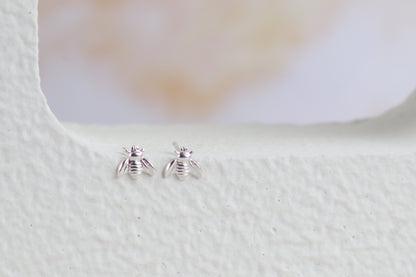 A pair of tiny silver bee stud earrings resting on a textured white surface with a soft, blurred light background.