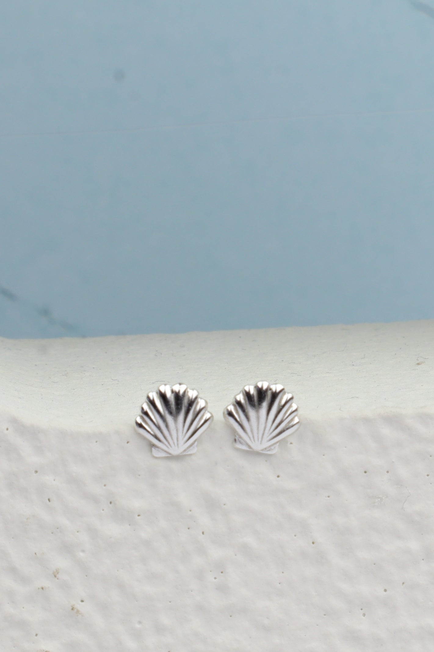 A pair of silver stud earrings shaped like small scallop shells, resting on a textured white ledge against a light blue background.