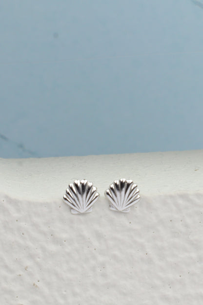 A pair of silver stud earrings shaped like small scallop shells, resting on a textured white ledge against a light blue background.
