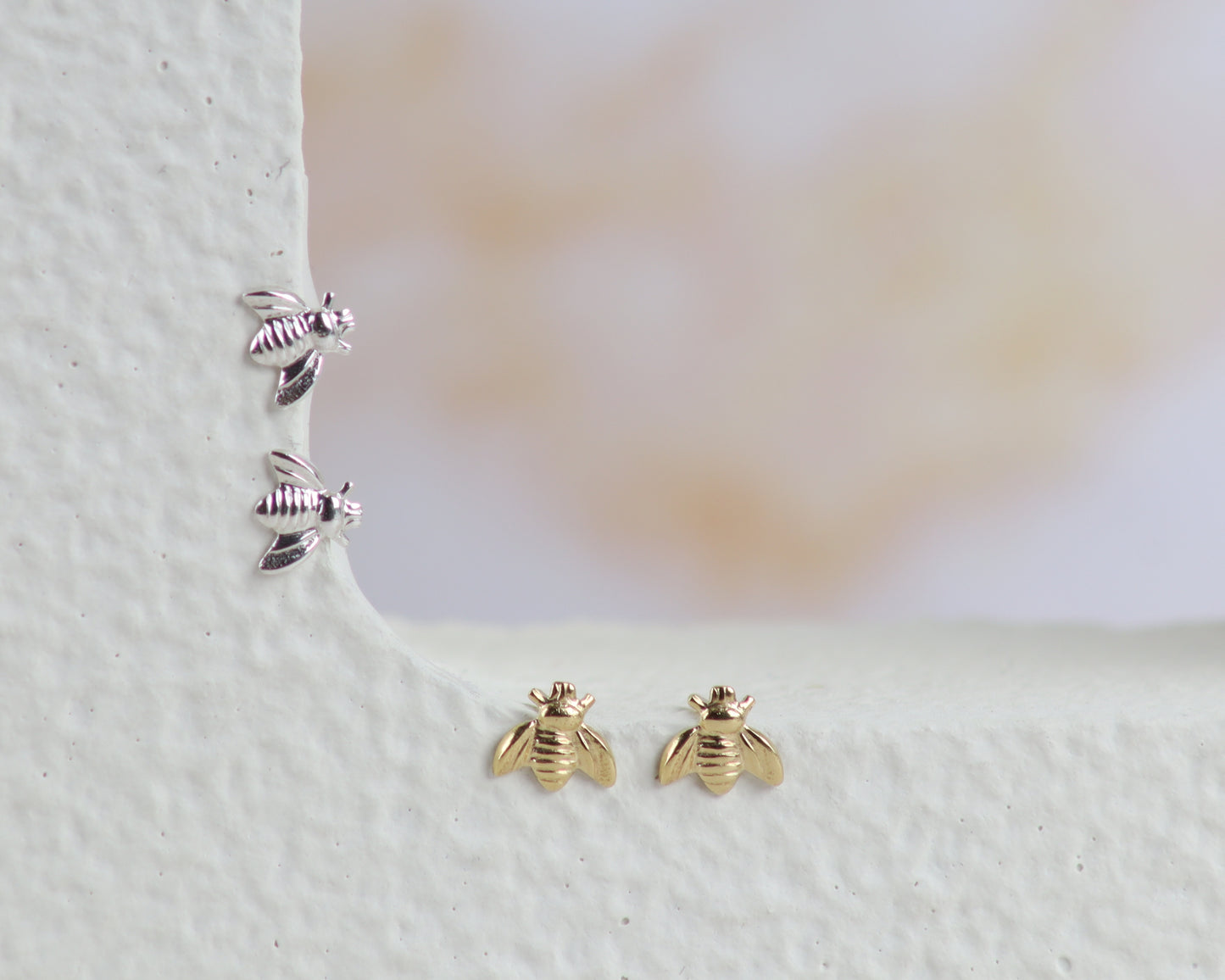A pair of gold bee stud earrings and a pair of silver bee stud earrings arranged on a textured white ledge against a soft, blurred beige background.