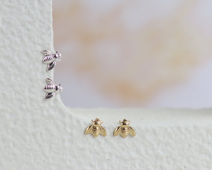 A pair of gold bee stud earrings and a pair of silver bee stud earrings arranged on a textured white ledge against a soft, blurred beige background.