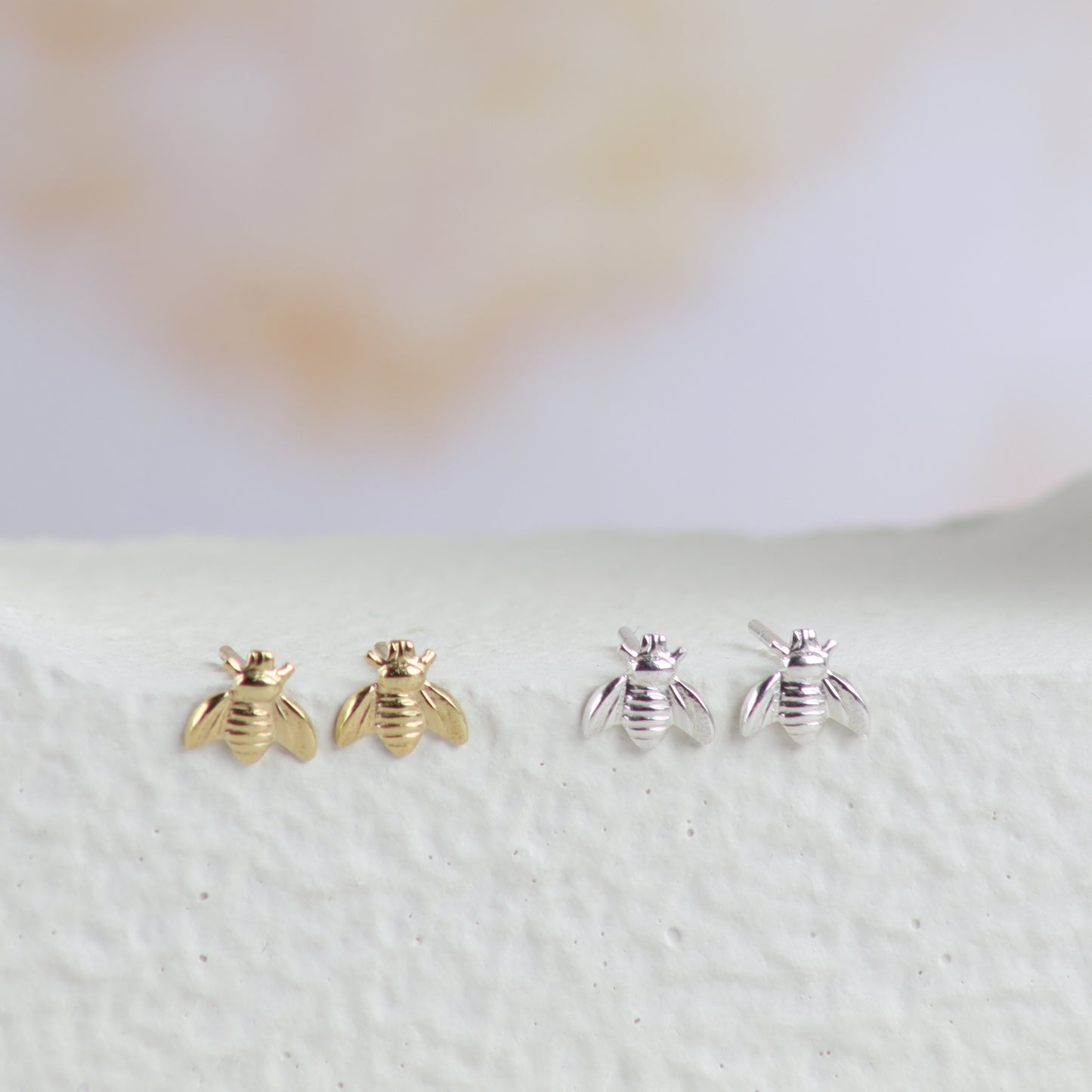 Three pairs of small bee-shaped stud earrings displayed on a textured white surface against a soft blurred light background. The left pair is gold-toned, and the right pair is silver-toned.