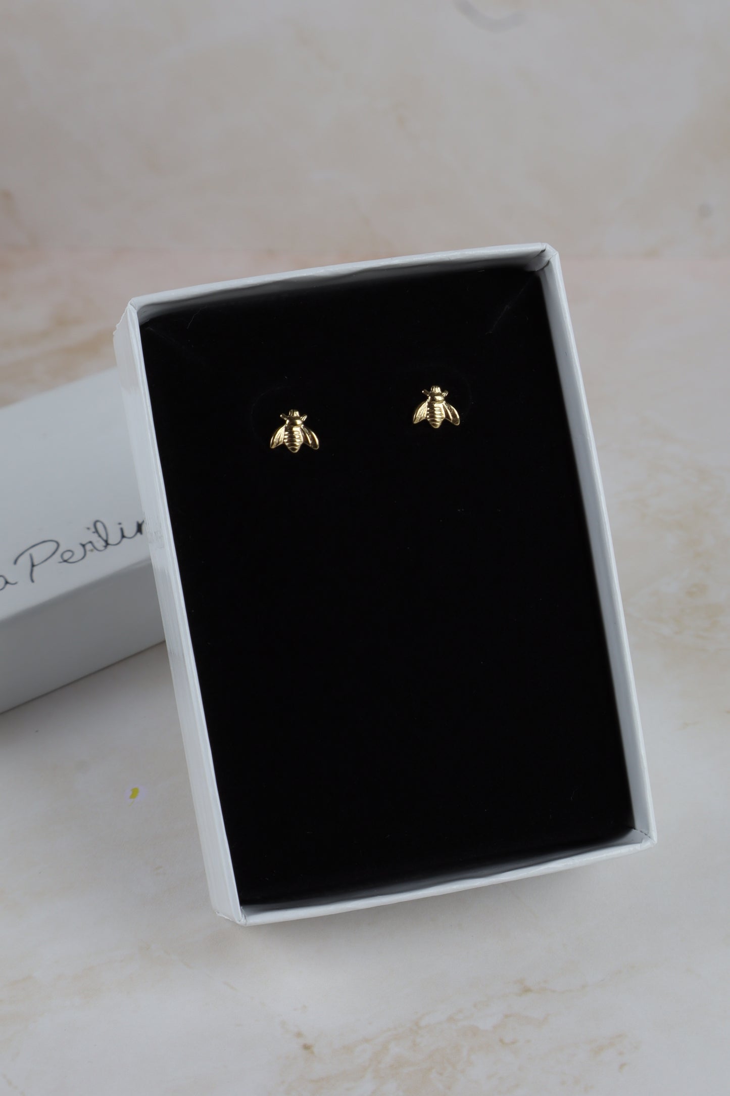 A pair of small gold bee stud earrings displayed on a black velvet insert inside an open white jewelry box, with part of a branded white box visible on the left.
