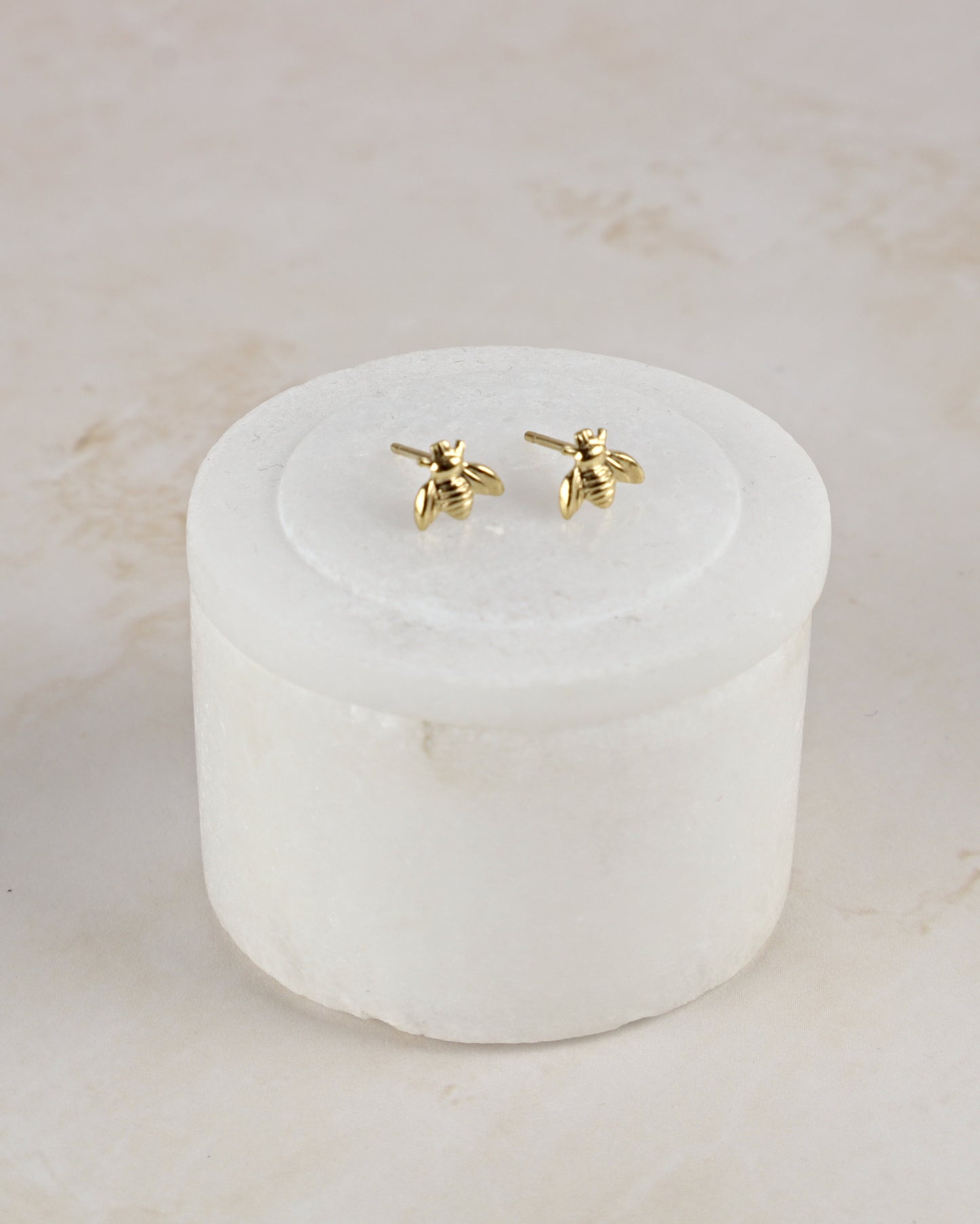 A pair of small gold bee stud earrings displayed on top of a round white stone holder against a light beige marble background.