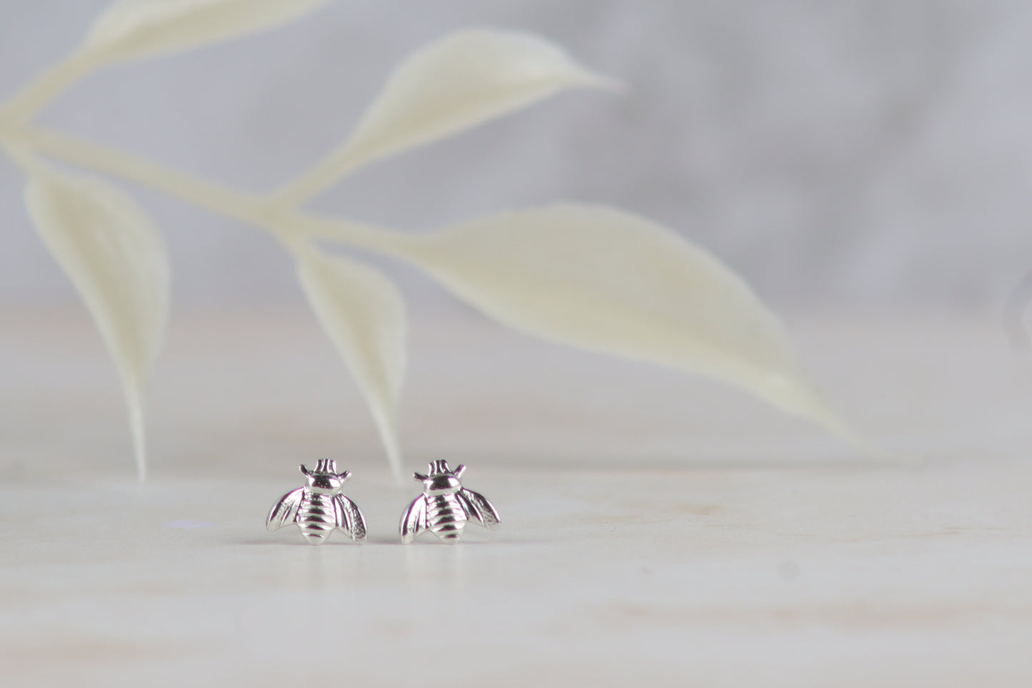 A pair of silver bee stud earrings resting on a light marble surface with a soft-focus leafy background.