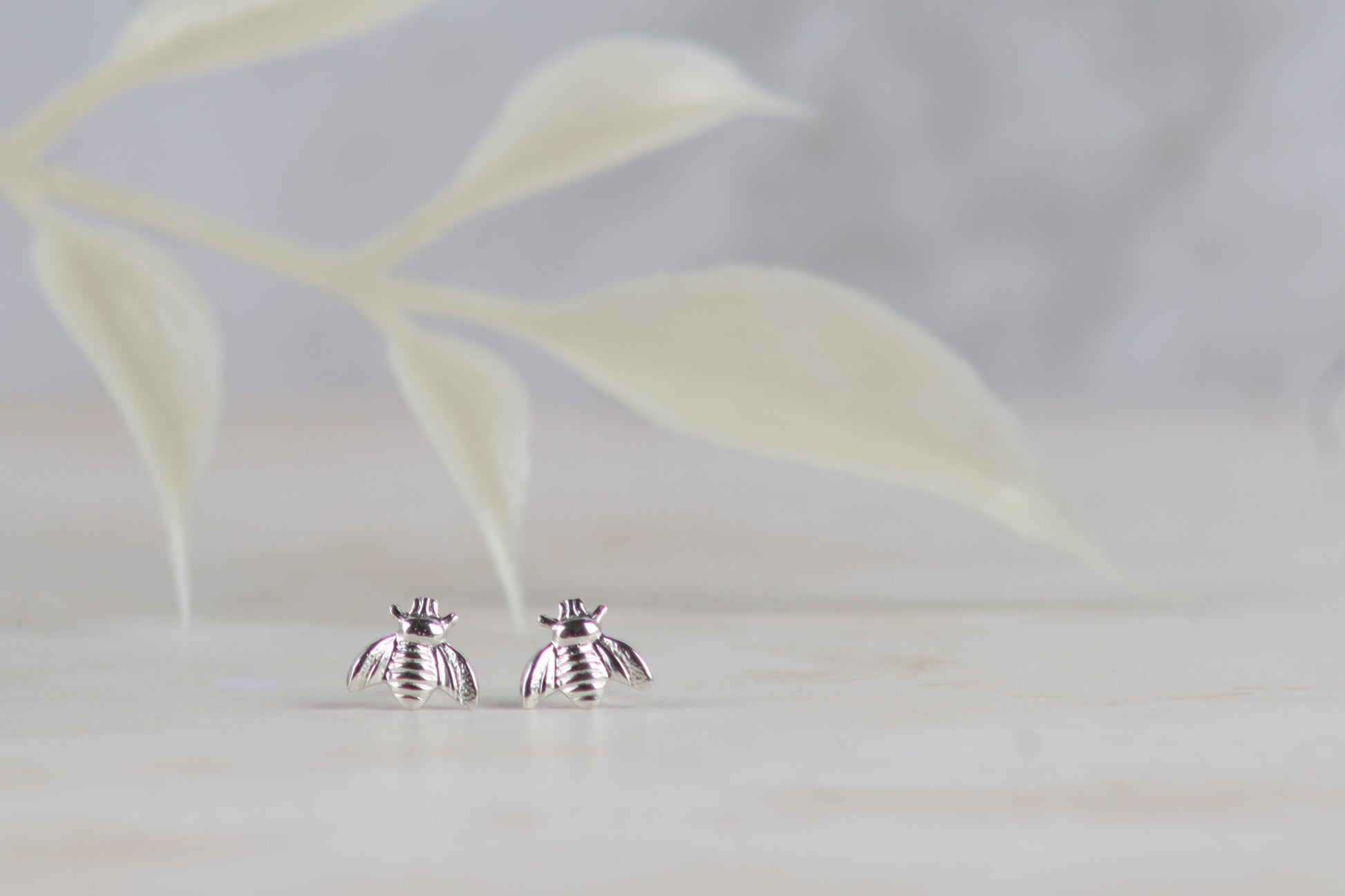 A pair of silver bee stud earrings resting on a light marble surface with a soft-focus leafy background.
