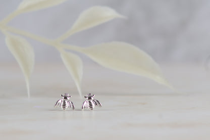 A pair of silver bee stud earrings resting on a light marble surface with a soft-focus leafy background.