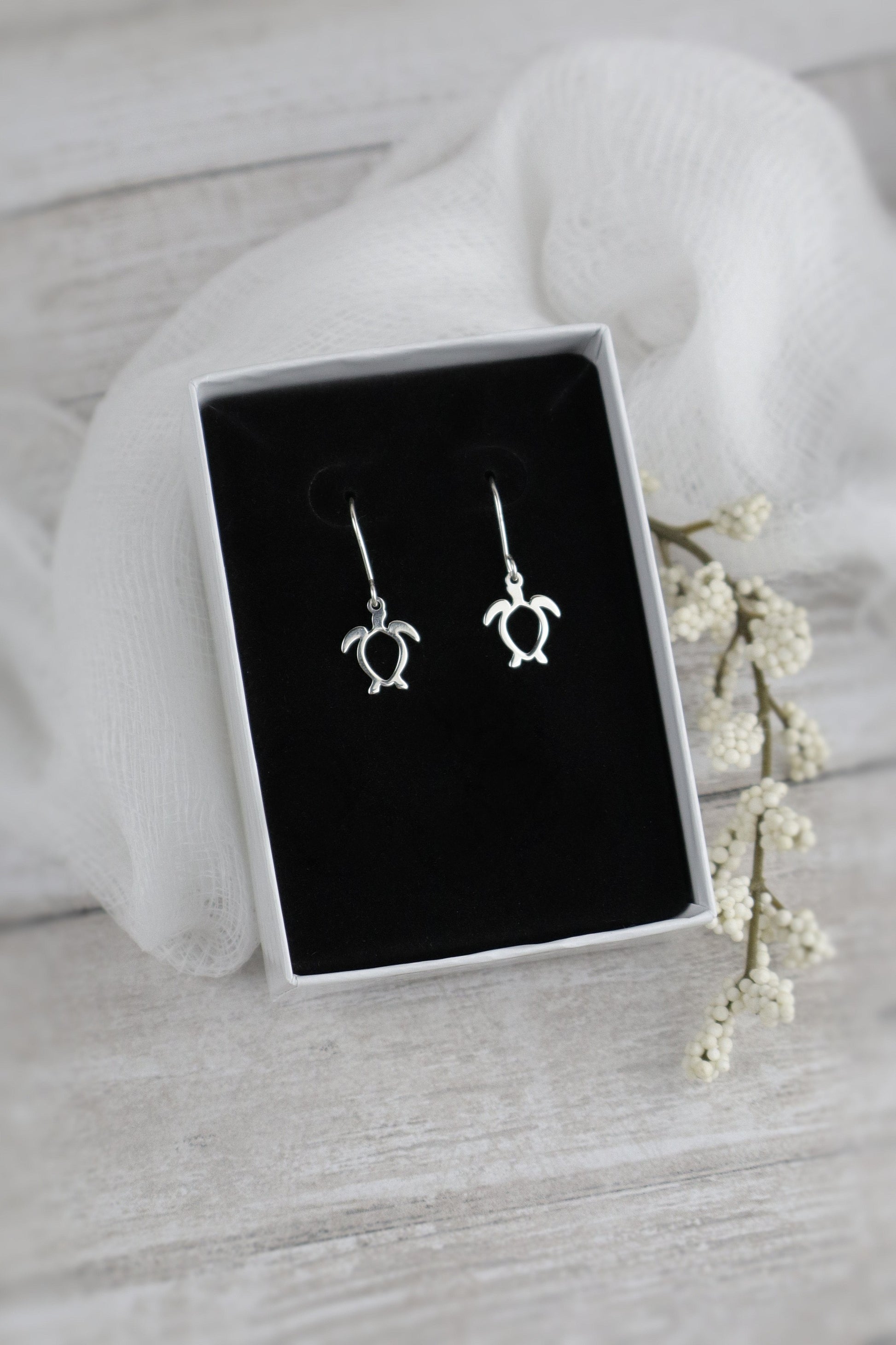Silver turtle-shaped earrings in a white box on a light gray surface with white flowers.